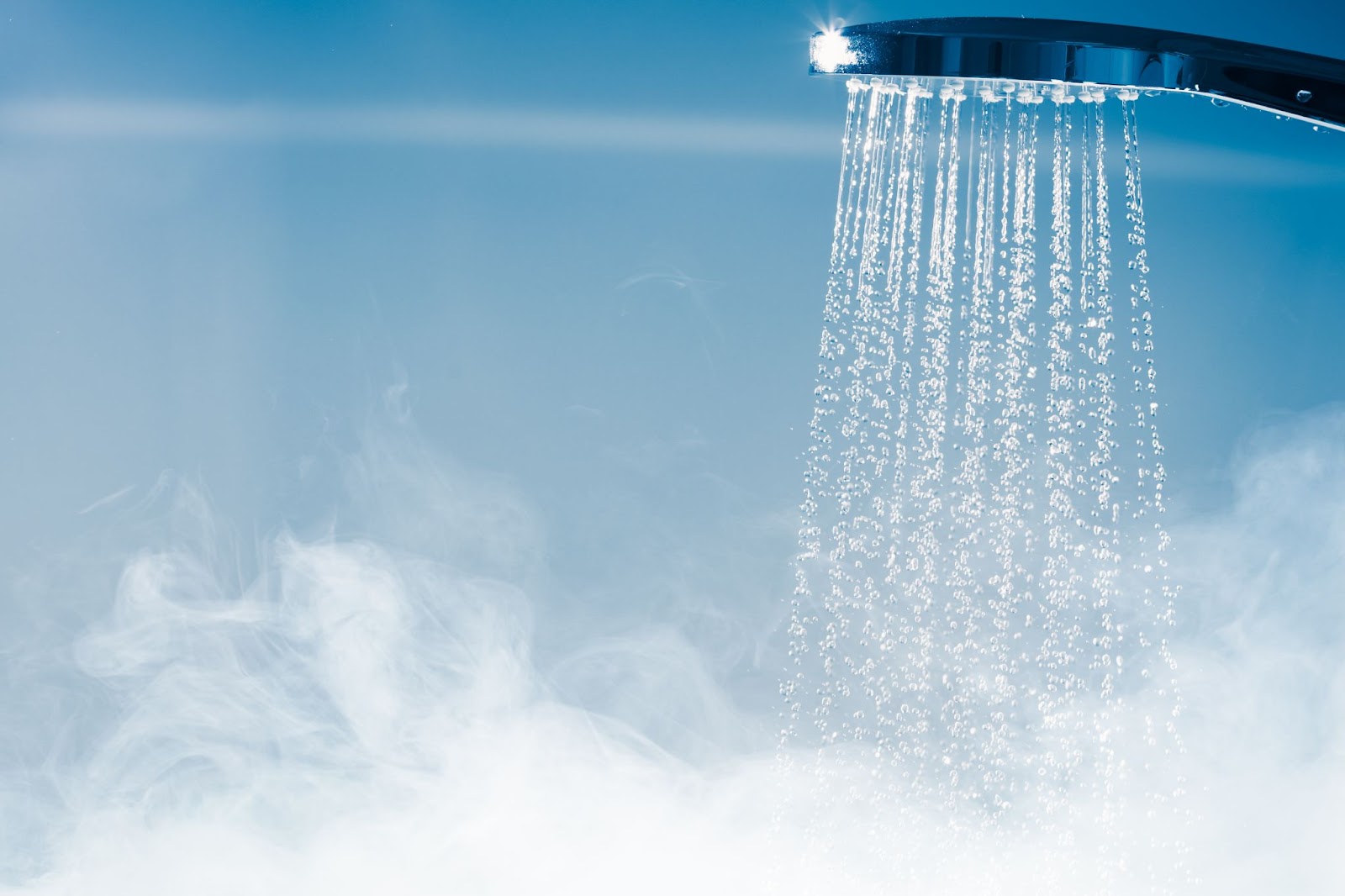 Low-angle view of a modern chrome shower head with water streaming down into a thick cloud of steam against a soft blue background.